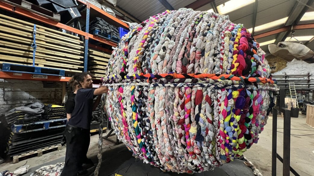A person stands beside a three-metre spherical sculpture made from colourful braided fabric and second-hand clothing, housed in an industrial warehouse with wooden pallets stacked behind.