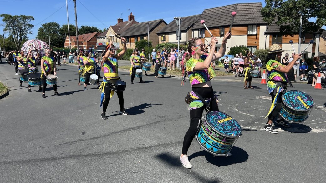 A samba drumming band in colourful neon costumes performs on a street, with a three-metre yarn ball made from multi-coloured plaited fabric visible in the background among spectators.