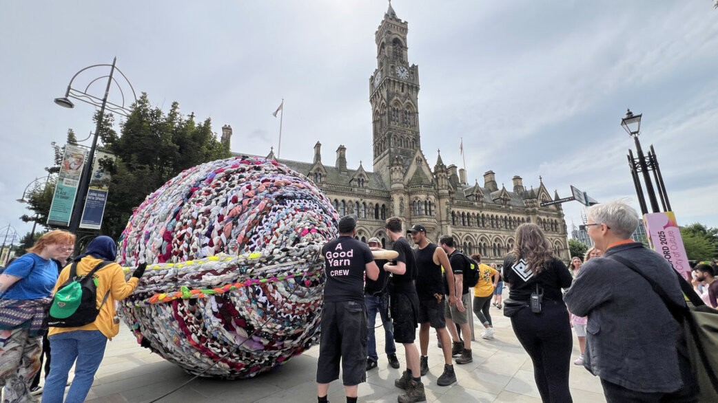 A giant yarn ball approximately three metres tall, made from colourful plaited fabric, surrounded by crew members and onlookers in front of Bradford City Hall.
