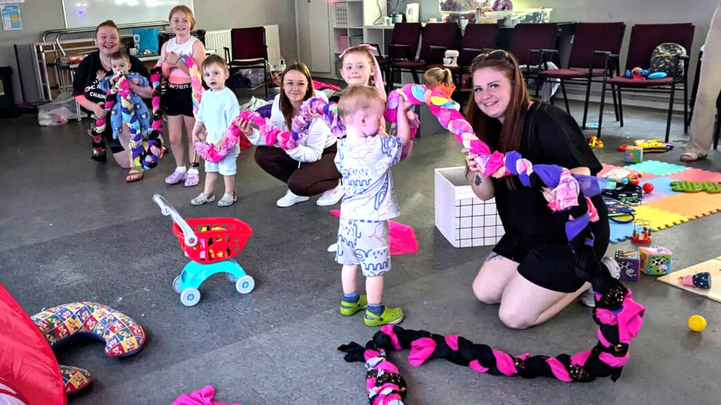 A group of adults and children hold up a colourful plaited rope made from pink, purple and black fabric during a community rope-making workshop in a hall.