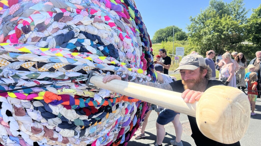 A bearded man in a cap pushes on a huge wooden knitting needle that's sticking out of a colourful three-metre yarn ball made from braided second-hand clothing, with onlookers watching on a sunny day.