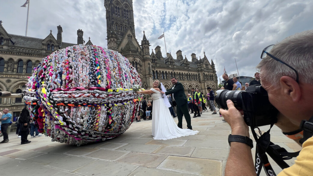 A bride and groom pose joyfully beside a three-metre-tall spherical sculpture made from plaited fabric ropes, while a photographer captures them in Bradford city centre.