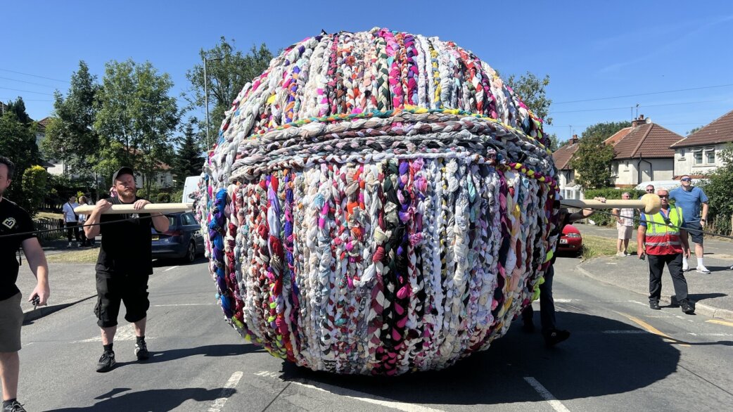 A colourful three-metre yarn ball made from braided second-hand clothing with a large knitting needle poking through it is rolled through a residential street by volunteers on a sunny day.