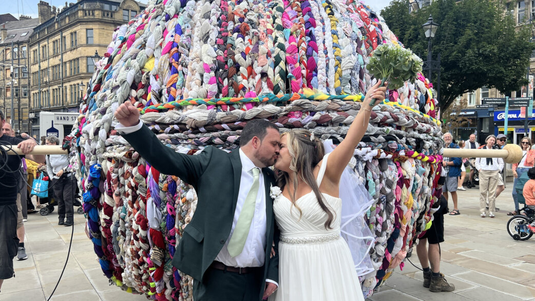 A bride and groom kiss with raised arms in front of a three-metre-tall yarn ball made from colourful braided fabric, displayed in a city square.