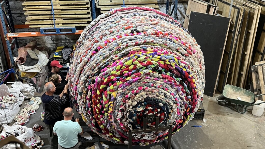 A giant multicoloured yarn ball, approximately three metres high, made from plaited second-hand clothing, sits in a warehouse as four people examine it from below.