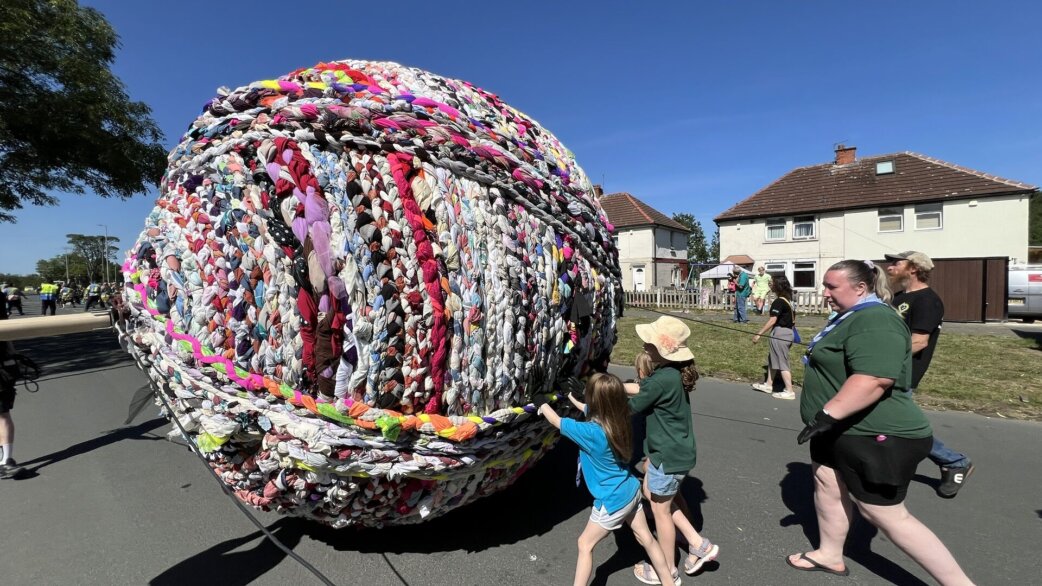 A giant colourful yarn ball, approximately three metres tall, made from plaited fabric and clothing, being rolled along a suburban street by children and adults on a sunny day.