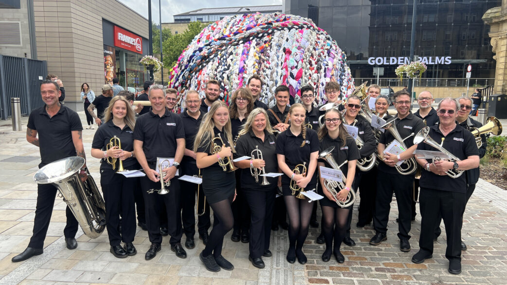 A brass band in matching black polo shirts poses in front of a three-metre spherical yarn ball made from colourful plaited fabric rope, in an urban square.