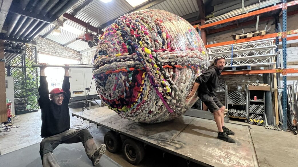 Two people pose beside a three-metre yarn ball made from plaited second-hand fabric and clothing, loaded on a flatbed trailer inside a warehouse. One of them is swinging from the giant knitting needle poking through the ball.