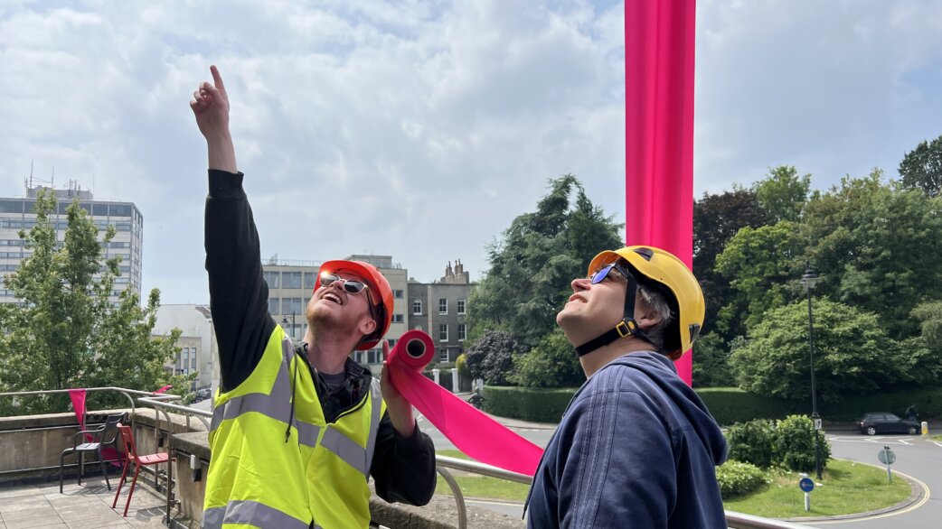 Two workers in hard hats and high-visibility clothing look upward at a bright magenta ribbon installation stretching vertically against a cloudy sky, with urban buildings and trees behind.