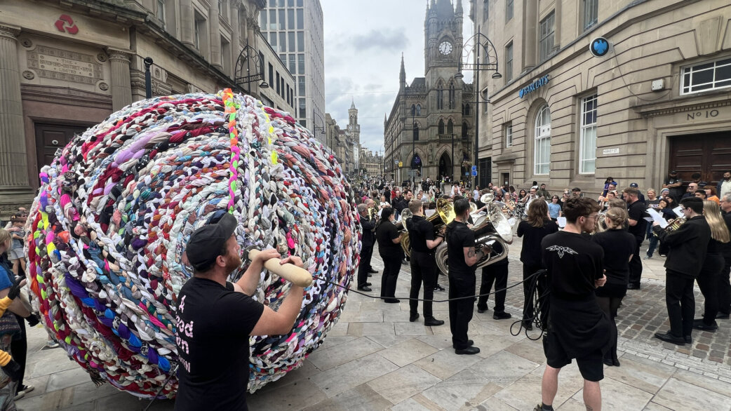 A giant three-metre yarn ball made from colourful braided fabric is rolled through a city street, accompanied by a brass band in black clothing and onlookers, with historic buildings in the background.