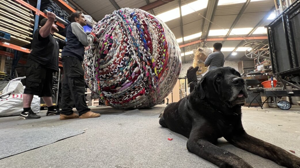 A large multicoloured yarn ball, approximately three metres high, made from braided fabric strips, being moved by workers in a warehouse. A black dog lies in the foreground.