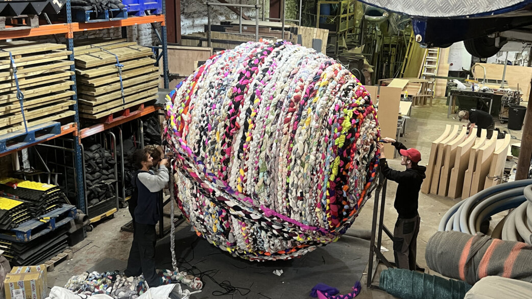 Two people position a colourful three-metre spherical sculpture made from plaited fabric rope in a warehouse workshop, with industrial shelving and equipment visible behind.