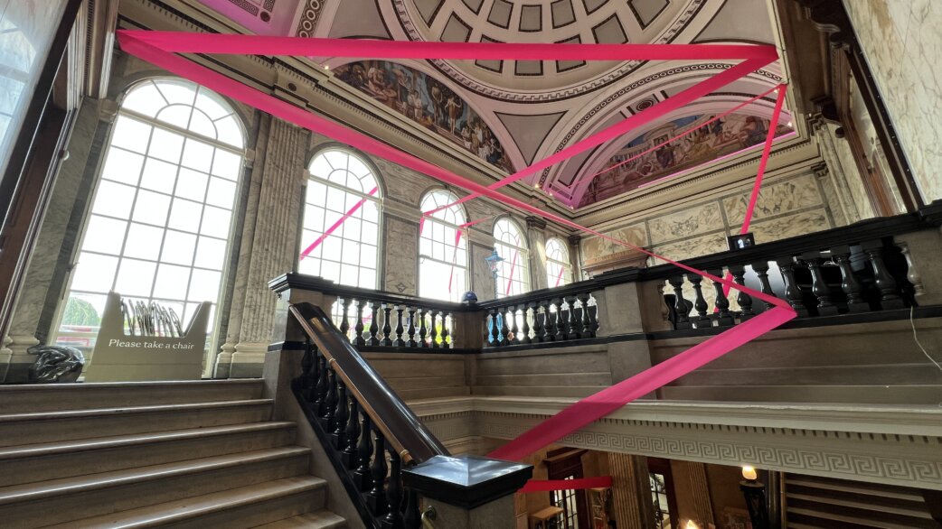 Bright magenta ribbon installation stretching diagonally across a grand Victorian stairwell with ornate domed ceiling, classical murals, and large arched windows at the Royal West of England Academy.