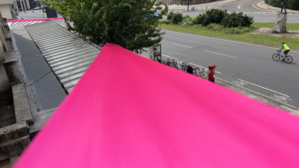 A bright magenta ribbon extends from an elevated viewpoint across steps and towards a street below, where a cyclist passes. Trees and parked bicycles visible in an urban setting.