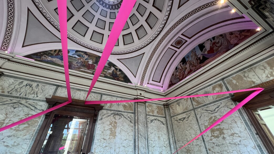 Bright magenta ribbons stretch diagonally across a grand classical interior with marble walls, ornate domed ceiling and painted friezes at the Royal West of England Academy.