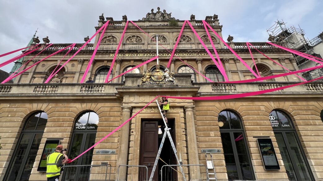 Workers on ladders install bright magenta ribbon across the stone façade of the Royal West of England Academy, creating a web-like pattern spanning approximately 15 metres across the historic building's front.