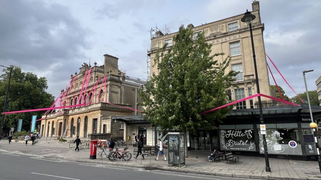 Bright magenta ribbon stretched across the exterior of the Royal West of England Academy and neighbouring buildings in Bristol, creating a linear sculptural installation against the stone architecture.