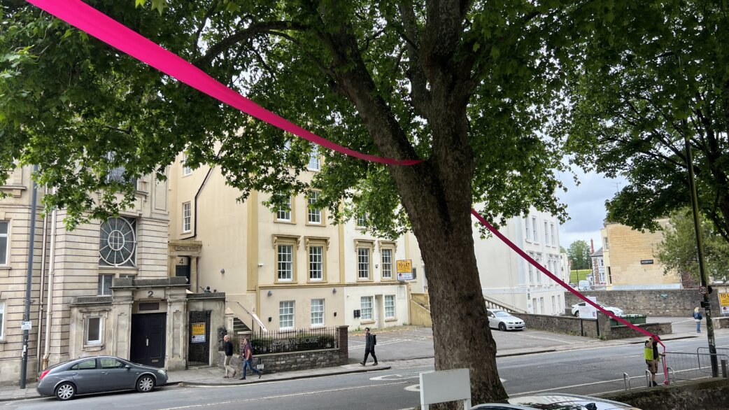 Bright magenta ribbon stretches across a Bristol street, wrapped around a large tree and extending between Georgian buildings including Beacon House, creating a striking linear intervention against the urban landscape.