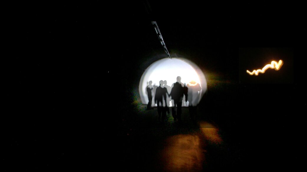 Silhouettes of several people walking through a circular tunnel opening, backlit by bright white light, with a wavy orange light trail visible to the right in the darkness.