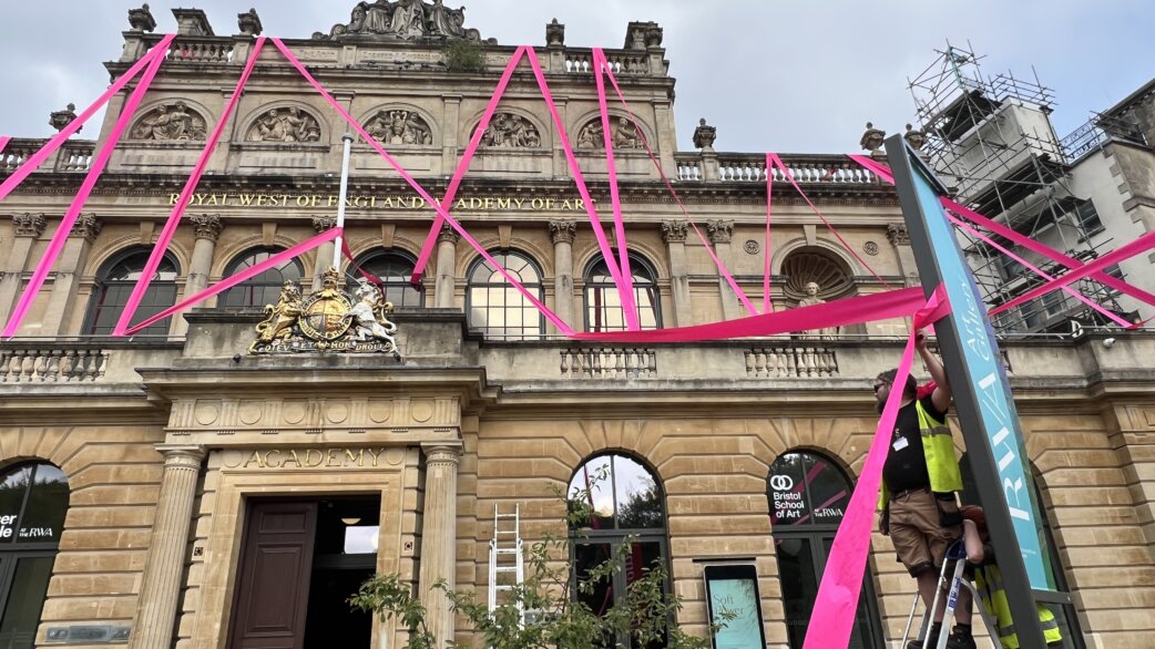 Bright magenta ribbons stretch across the classical stone facade of the Royal West of England Academy, with workers on ladders securing the installation to a signpost.
