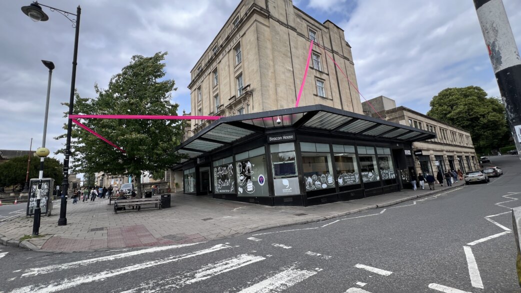 A bright magenta ribbon stretches diagonally across an urban street corner, connecting Beacon House to nearby buildings. The ribbon forms part of a 1km experimental line drawing installation in Bristol.
