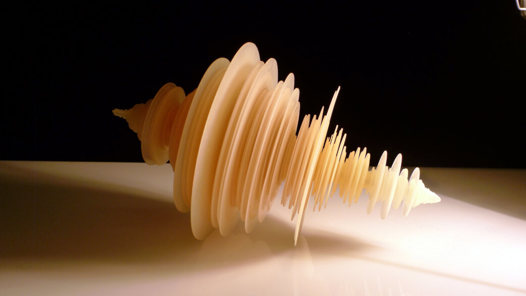 A cream-coloured 3D-printed sculpture resembling a rotating seismogram waveform, with varying disc-like rings creating an irregular spindle shape, approximately 30cm long, against a black background.