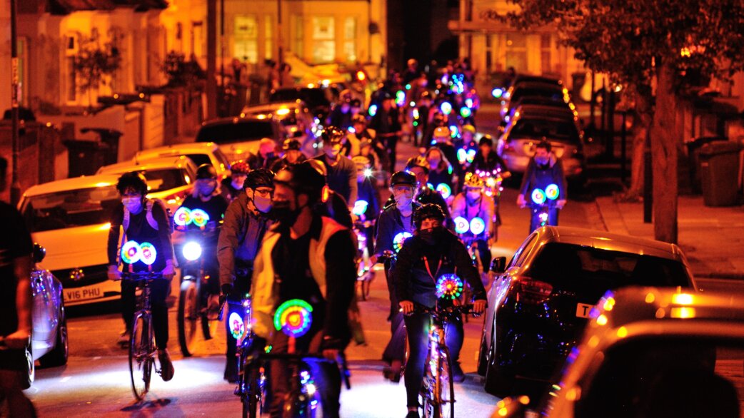 A nighttime street scene showing dozens of cyclists riding with colourful LED wheel lights creating circular rainbow patterns, illuminating a residential road in warm orange tones.