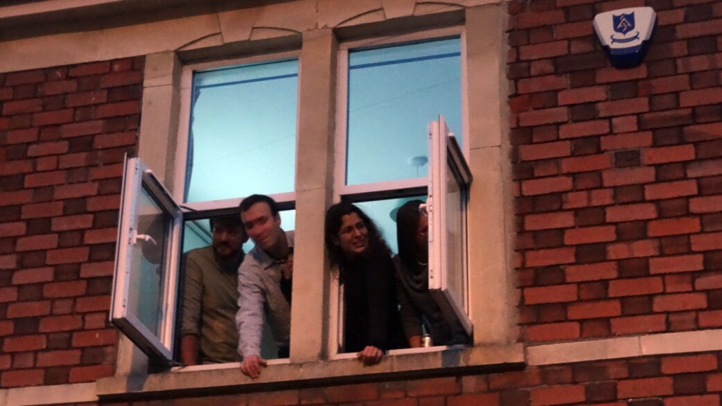 Four people lean out of open windows in a brick building at night, smiling and looking down at something below, likely watching a street performance.