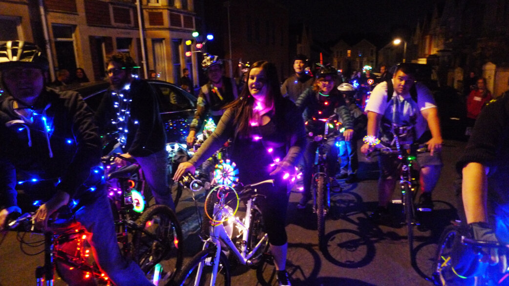 A group of cyclists ride at night on a residential street, their bicycles decorated with colourful LED lights in blue, purple, red and multicoloured strands.