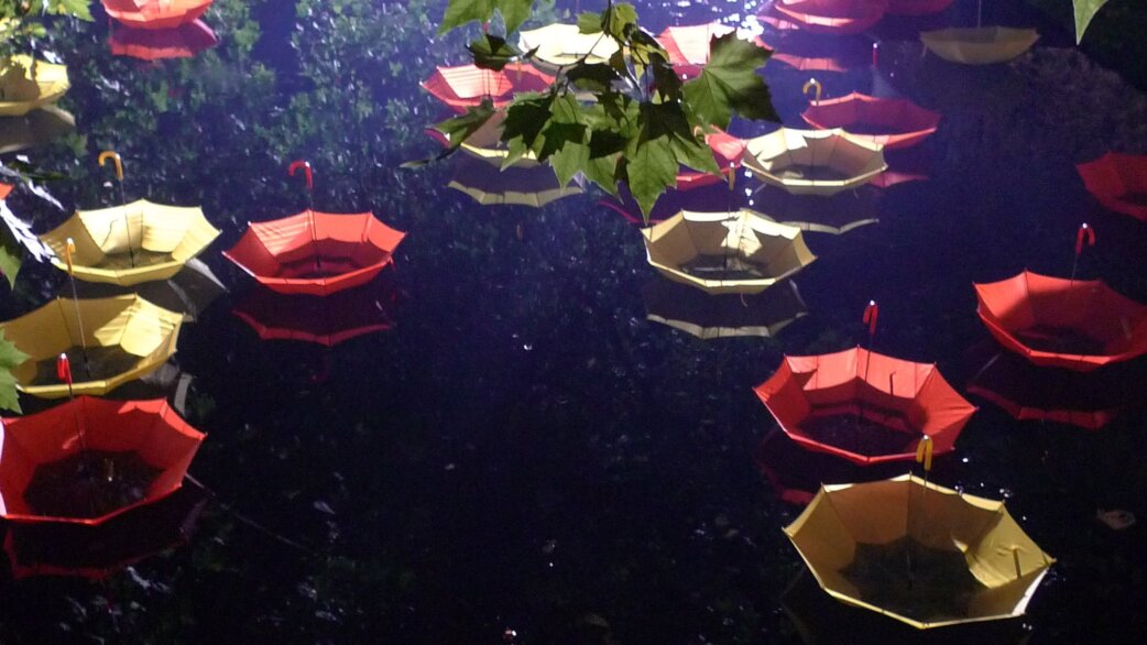 Colourful umbrellas in red, yellow and orange floating upside-down on dark canal water at night, with green foliage visible above, creating a whimsical reflection.