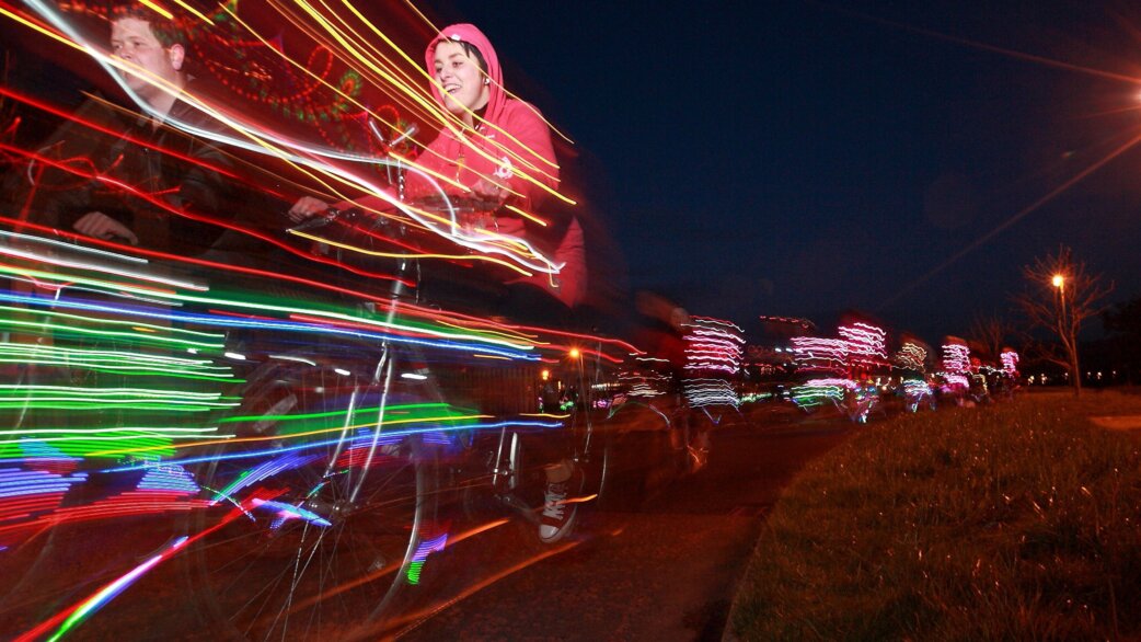 Cyclists riding at night with colourful LED lights attached to their bikes, creating streaks of yellow, green, blue and red light trails through long exposure photography.
