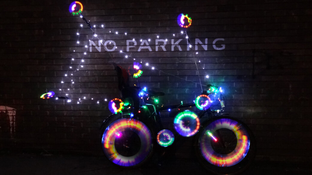 A bicycle decorated with colourful LED lights on its wheels and frame, parked at night against a brick wall with "NO PARKING" painted in white letters.
