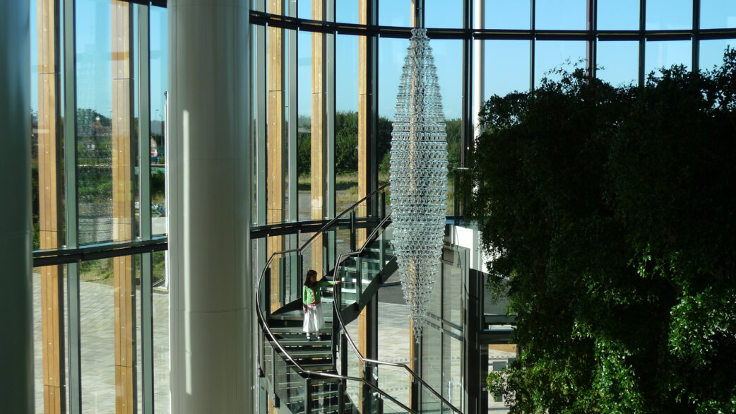 A tall conical chandelier made of hundreds of glass radiometers, approximately 5 metres high, suspended in a modern glass atrium with a spiral staircase and a figure for scale.