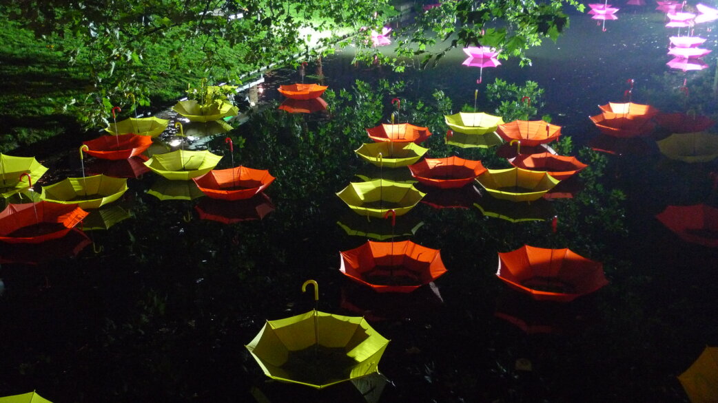 Hundreds of upturned yellow, orange and pink umbrellas floating on a canal at night, illuminated against dark water and surrounded by green foliage.