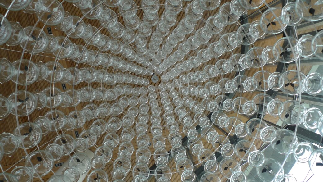 A kinetic chandelier viewed from below, comprising hundreds of glass radiometers arranged in concentric circles, suspended against a wooden slatted ceiling.