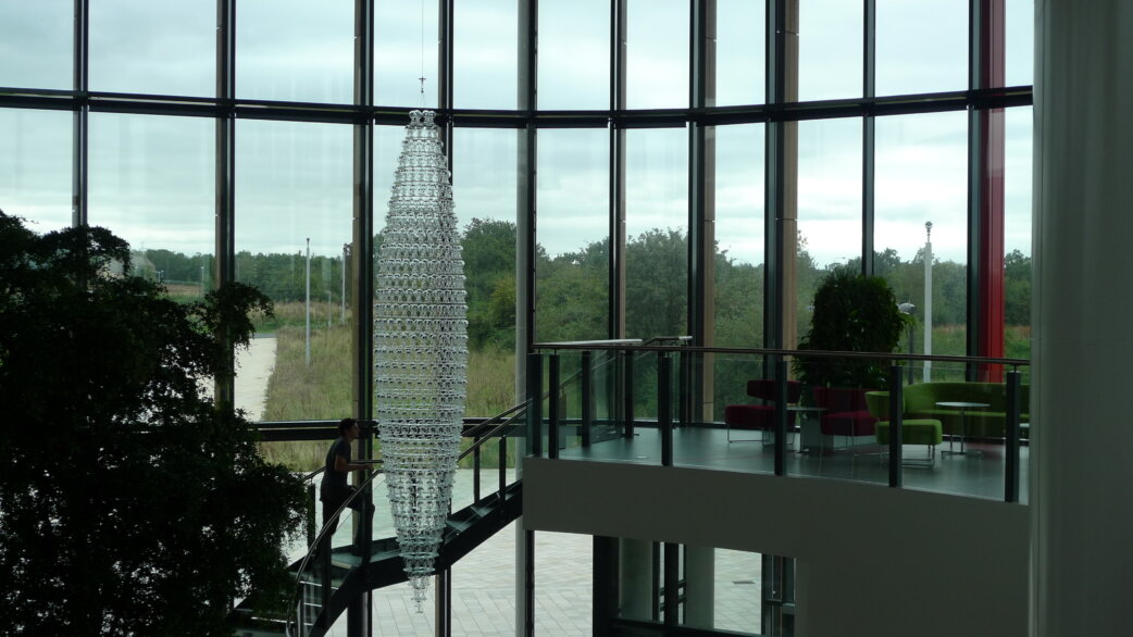 A tall conical chandelier made of hundreds of glass radiometers, approximately 5 metres high, suspended in a modern atrium with floor-to-ceiling windows overlooking parkland.