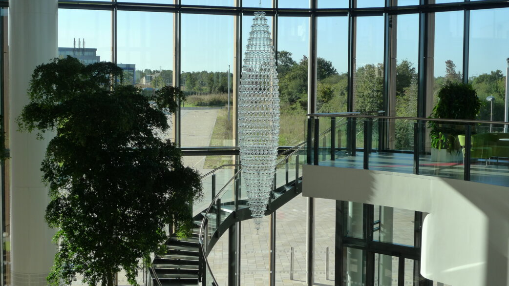 A tall conical chandelier made of hundreds of glass radiometers hangs in a modern glass-walled atrium, approximately 5 metres tall, with a curved staircase and indoor trees nearby.