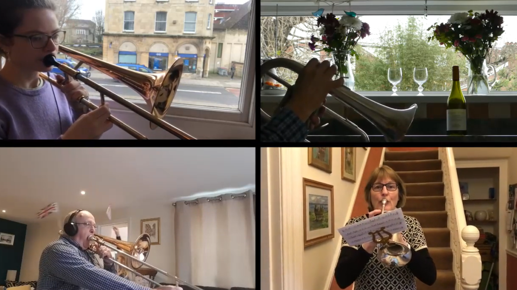 Four musicians playing brass instruments from their homes in a video call grid layout, performing trombones and a cornet as part of a remote ensemble during lockdown.