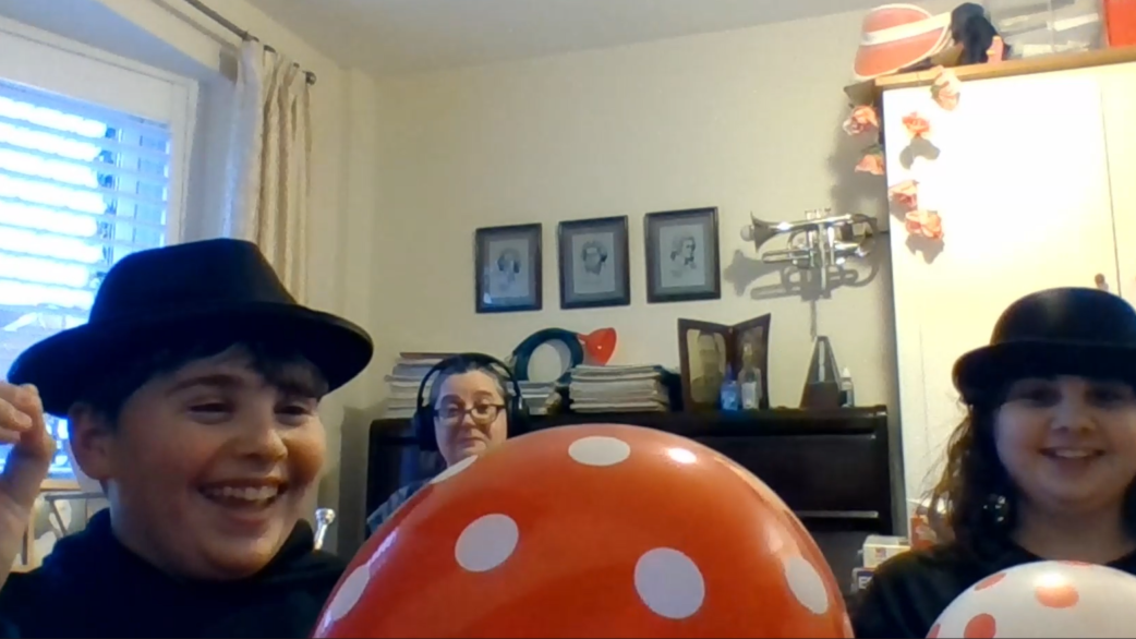 Two children wearing black bowler hats smile while holding orange polka-dot balloons, with an adult wearing headphones seated at a piano behind them in a home setting.