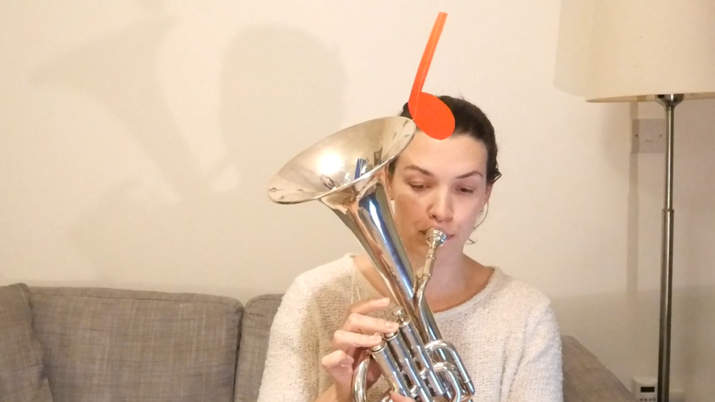 A woman in a white jumper plays a silver tenor horn while seated on a sofa, with an orange musical note prop attached to the instrument's bell.