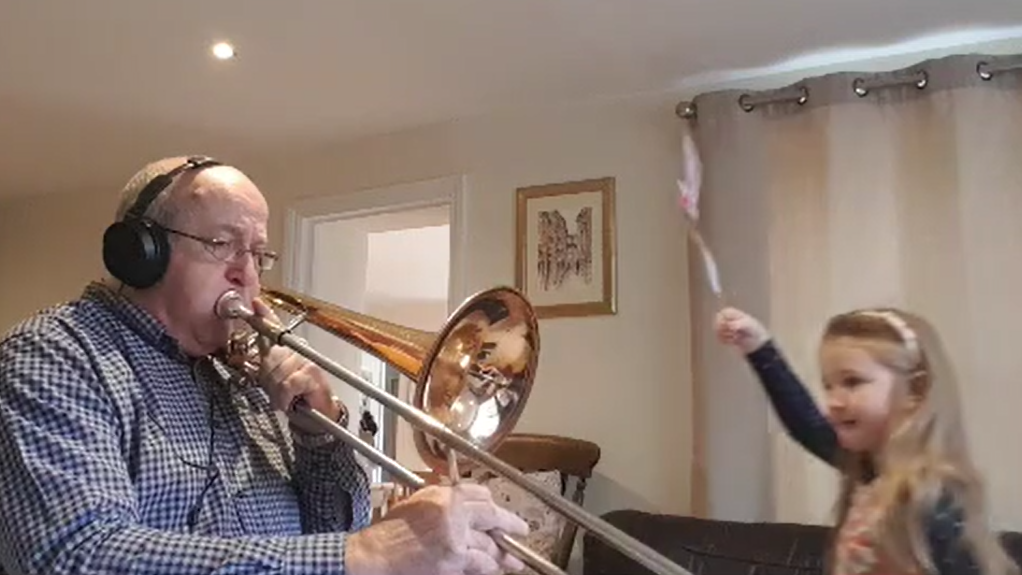 An older man wearing headphones plays a trombone while a young girl waves a wand enthusiastically beside him, filmed in a home setting for a reversed music project.