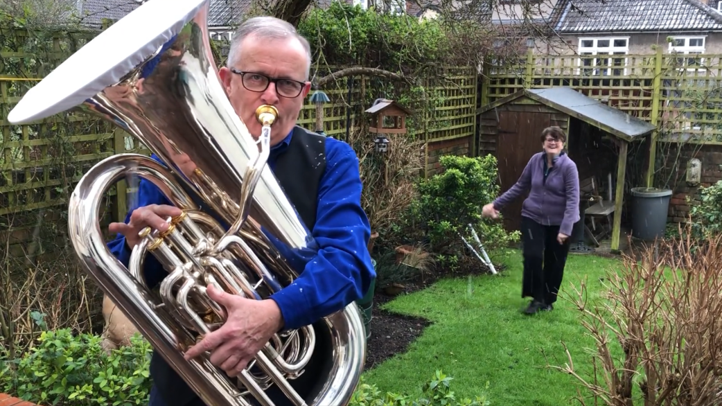 A man in a blue shirt plays a silver euphonium in a suburban back garden while a woman stands watching in the background near a wooden shed.