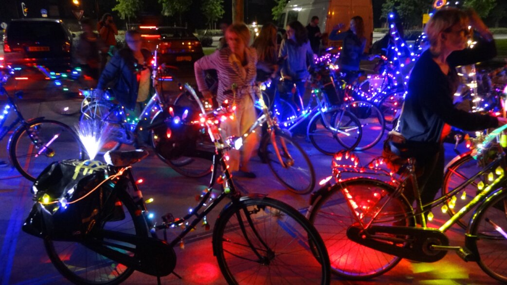 Nighttime scene of cyclists gathered with bicycles adorned in colourful LED fairy lights, creating a festive, glowing atmosphere on an urban street.