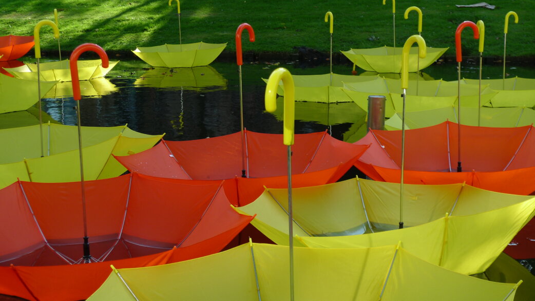 Brightly coloured yellow and orange umbrellas floating upside-down on a canal, their curved handles pointing upward, with green grass visible in the background.